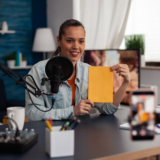 Woman influencer presenting yellow book in front of recording smartphone. Similing content creator broadcasting review for social media channel in home studio with microphone and laptop.