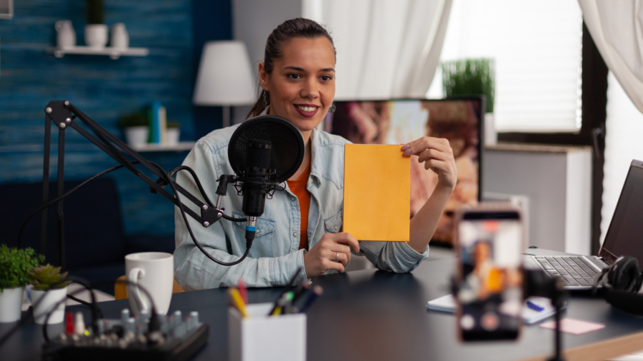 Woman influencer presenting yellow book in front of recording smartphone. Similing content creator broadcasting review for social media channel in home studio with microphone and laptop.